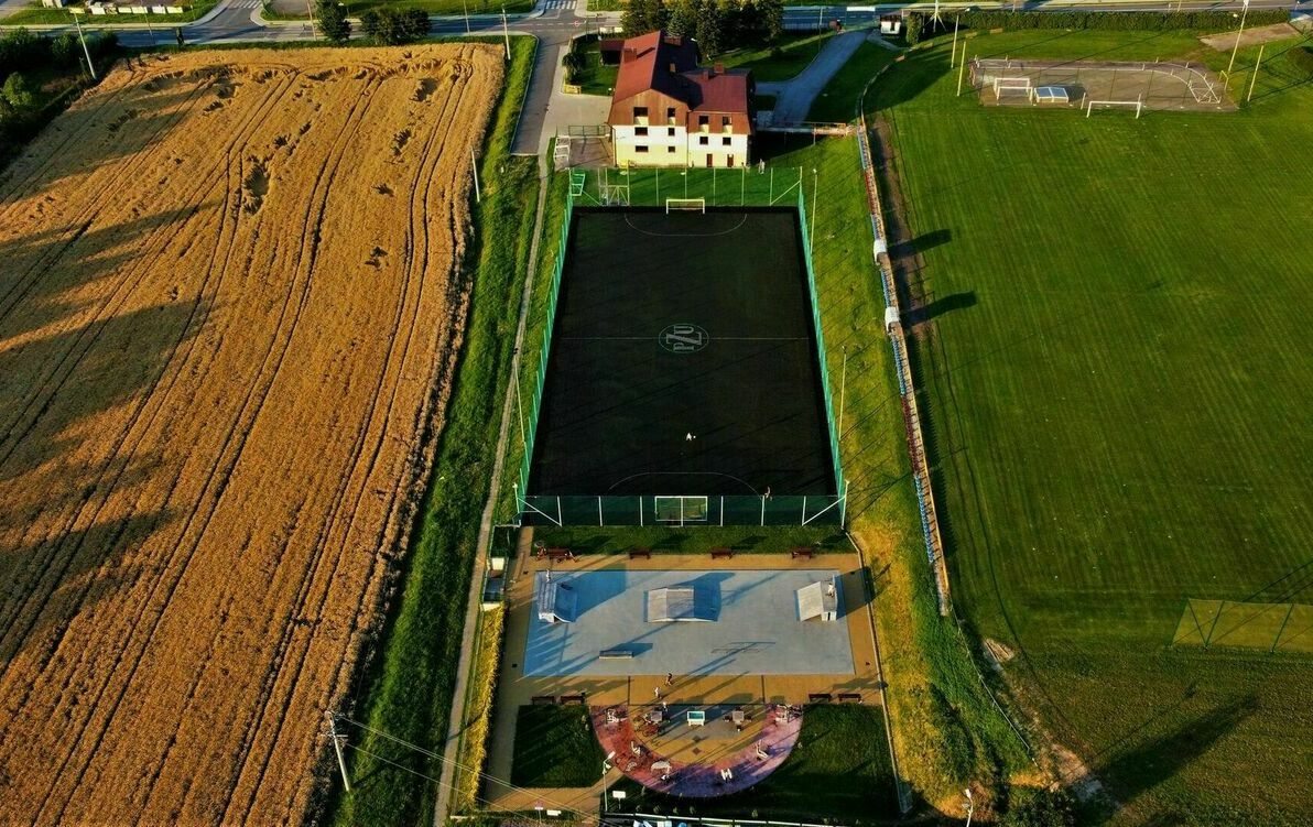 widok z góry na boisko sportowe ze sztuczną trawą, powyżej boiska znajduje się budynek  w stulu PRL , po prawej stronie boiska jest stadion a po lewej  pole uprawne