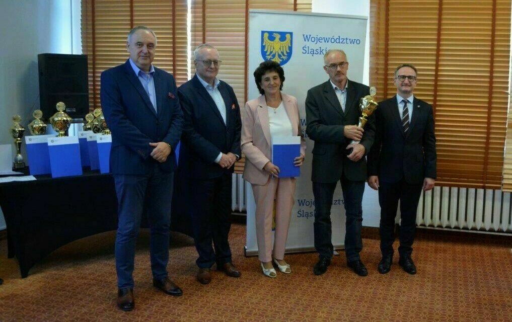 4 mężczyzn w garniturach i jedna kobieta w różowym eleganckim garniturze. Wszyscy w średnim wieku. w tle ścianka reklamująca województwo śląsikie i zestaw pucharów