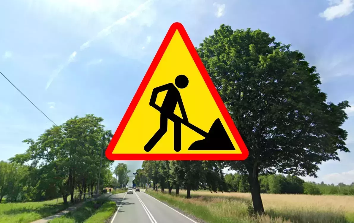 Znak drogowy „roboty drogowe” na tle wiejskiej drogi otoczonej drzewami i polami w słoneczny dzień.