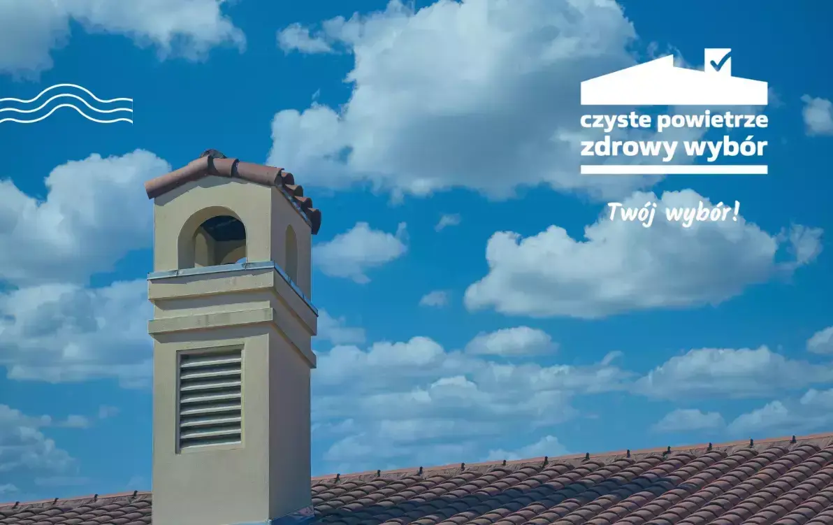 Kominek na dachu budynku z czerwonej dachówki na tle nieba z białymi chmurami. Logo programu Czyste Powietrze z hasłem „czyste powietrze zdrowy wybór. Twój wybór!” w prawym górnym rogu.