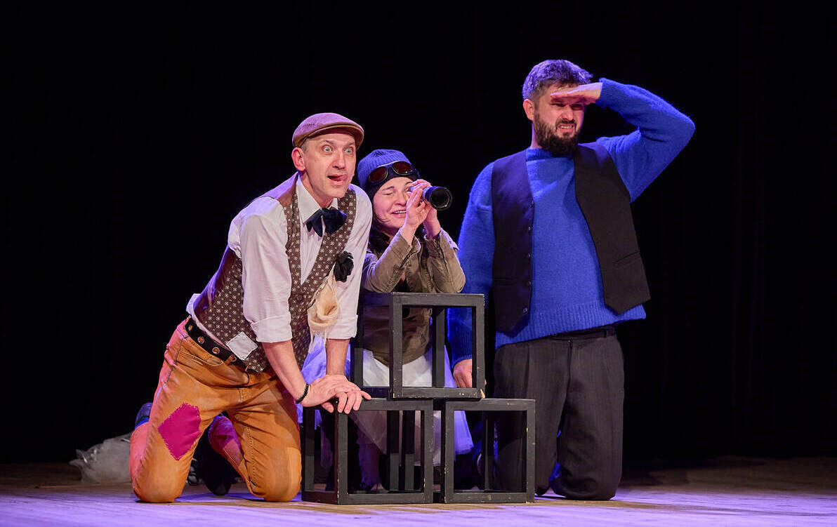 Trójka aktorów podczas spektaklu teatralnego na scenie. Dwóch mężczyzn i kobieta klęczą przy geometrycznej konstrukcji z czarnych ram. Jedna osoba patrzy przez lornetkę, druga osłania oczy dłonią jakby wypatrywała w oddali, trzecia pochyla się z zaciekawi