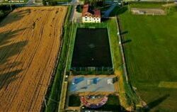 widok z góry na boisko sportowe ze sztuczną trawą, powyżej boiska znajduje się budynek  w stulu PRL , po prawej stronie boiska jest stadion a po lewej  pole uprawne