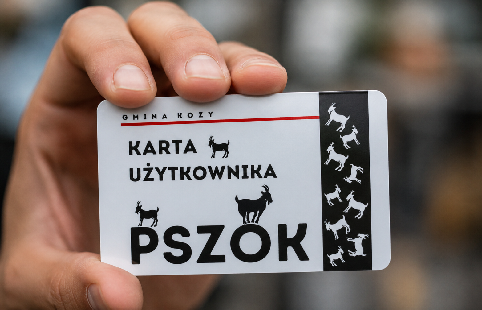 Dłoń trzyma realistyczną kartę użytkownika PSZOK Gminy Kozy na tle rozmytego otoczenia. Biała karta z czarnym napisem „KARTA UŻYTKOWNIKA PSZOK” ozdobiona jest grafikami kozłów oraz pionowym czarnym paskiem po prawej stronie. Zdjęcie wykonano w zbliżeniu, z naturalnym oświetleniem i małą głębią ostrości, co podkreśla profesjonalny wygląd projektu i nowoczesny charakter identyfikatora.